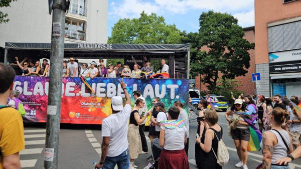 Der kunterbunte CSD-Truck 2025 mit feiernden Menschen. Davor ein Banner mit 'Glaube. Liebe. Laut.'